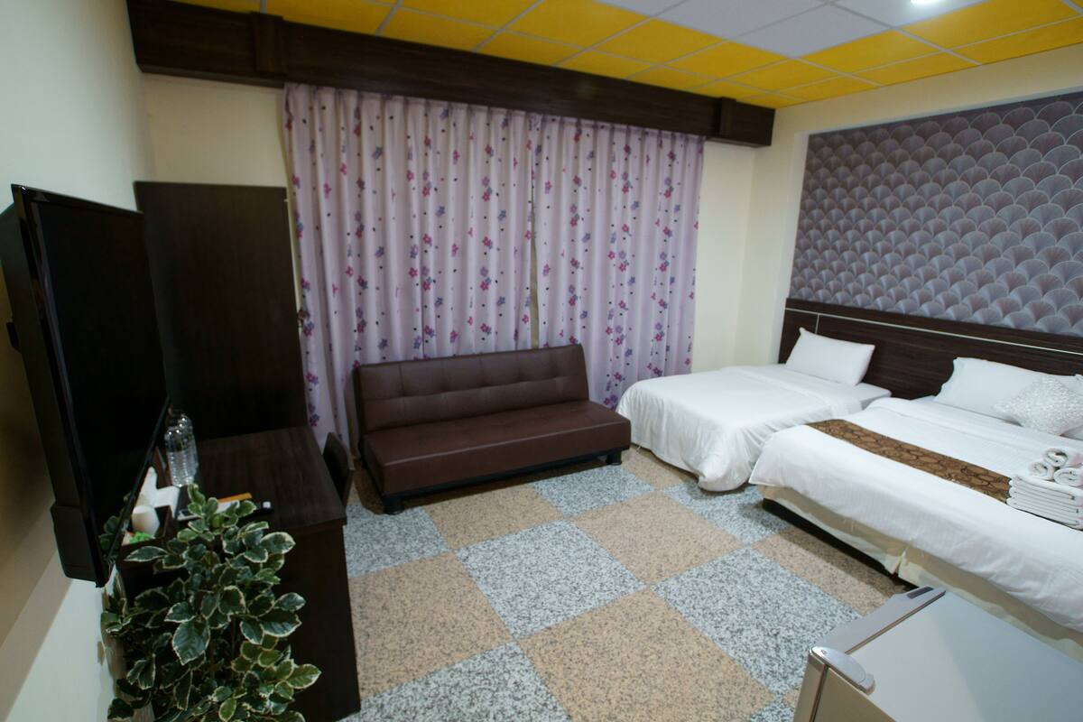 A hotel room with a sofa, two beds, a television, and a plant.