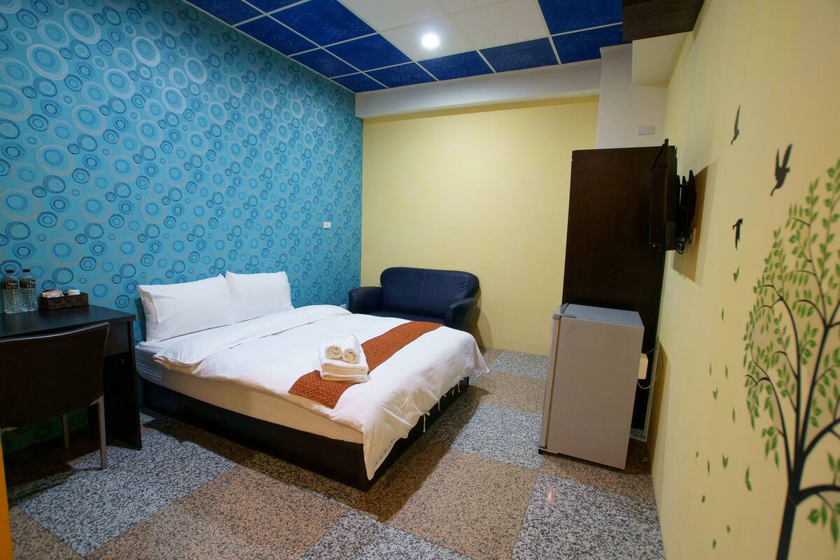 A hotel room with a bed, a desk, a chair, a refrigerator, and a wall with a tree mural.