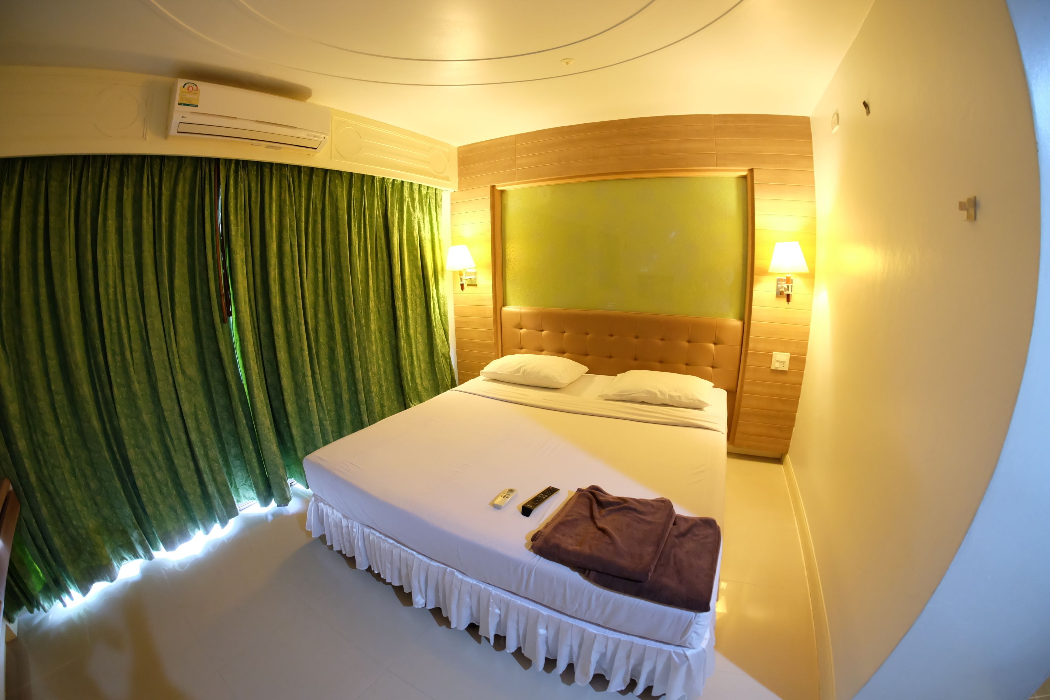 A hotel room with a large bed, green curtains, two wall-mounted lamps, and an air conditioning unit.