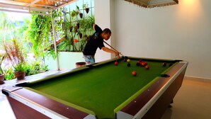 Une table de billard avec des billes, une personne qui joue, et des plantes en pot en arrière-plan.