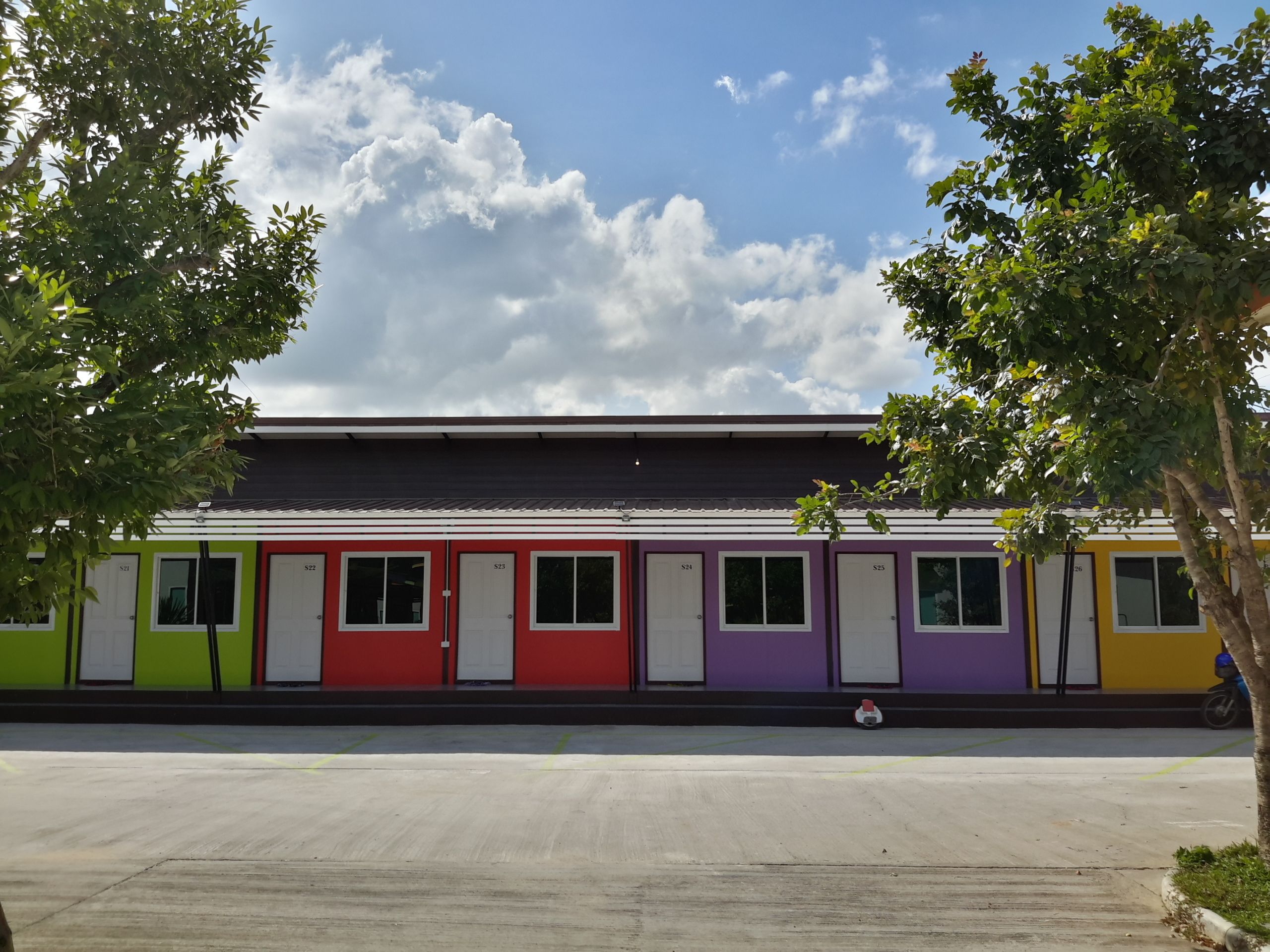 A row of colorful single-room cabins with individual doors, numbered from S21 to S26, set against a clear sky with clouds.