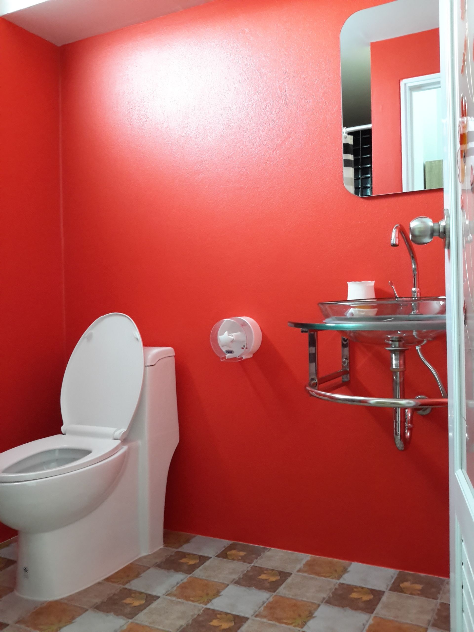 Une salle de bain avec un mur rouge, des toilettes blanches, un lavabo en verre avec un robinet chromé et un miroir au-dessus du lavabo.
