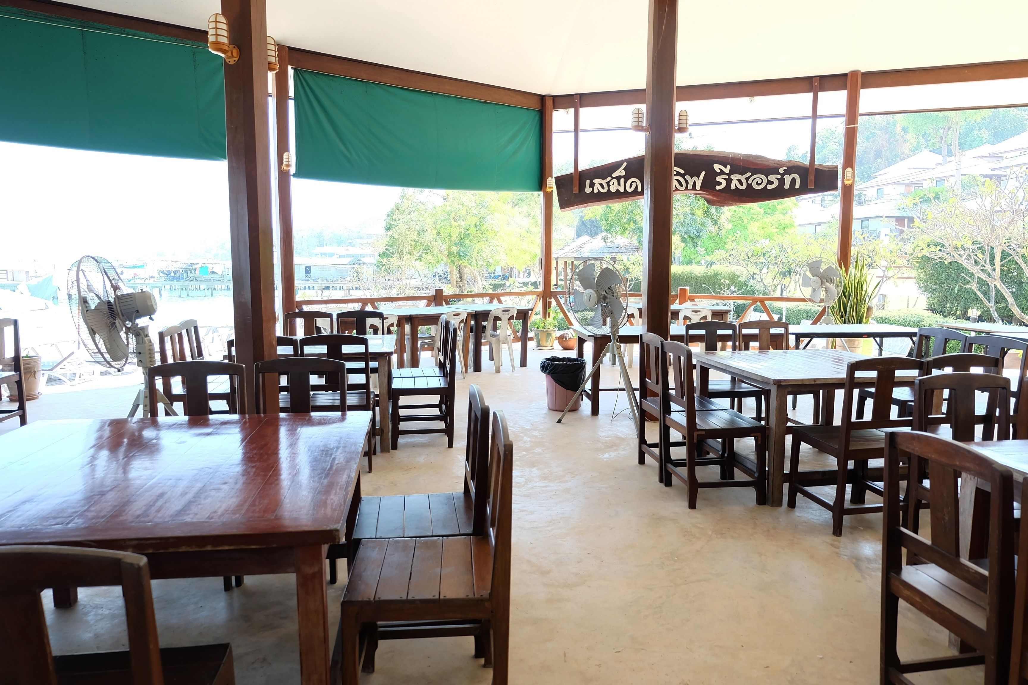 Un restaurante al aire libre con mesas y sillas de madera, ventanas amplias y un toldo verde.