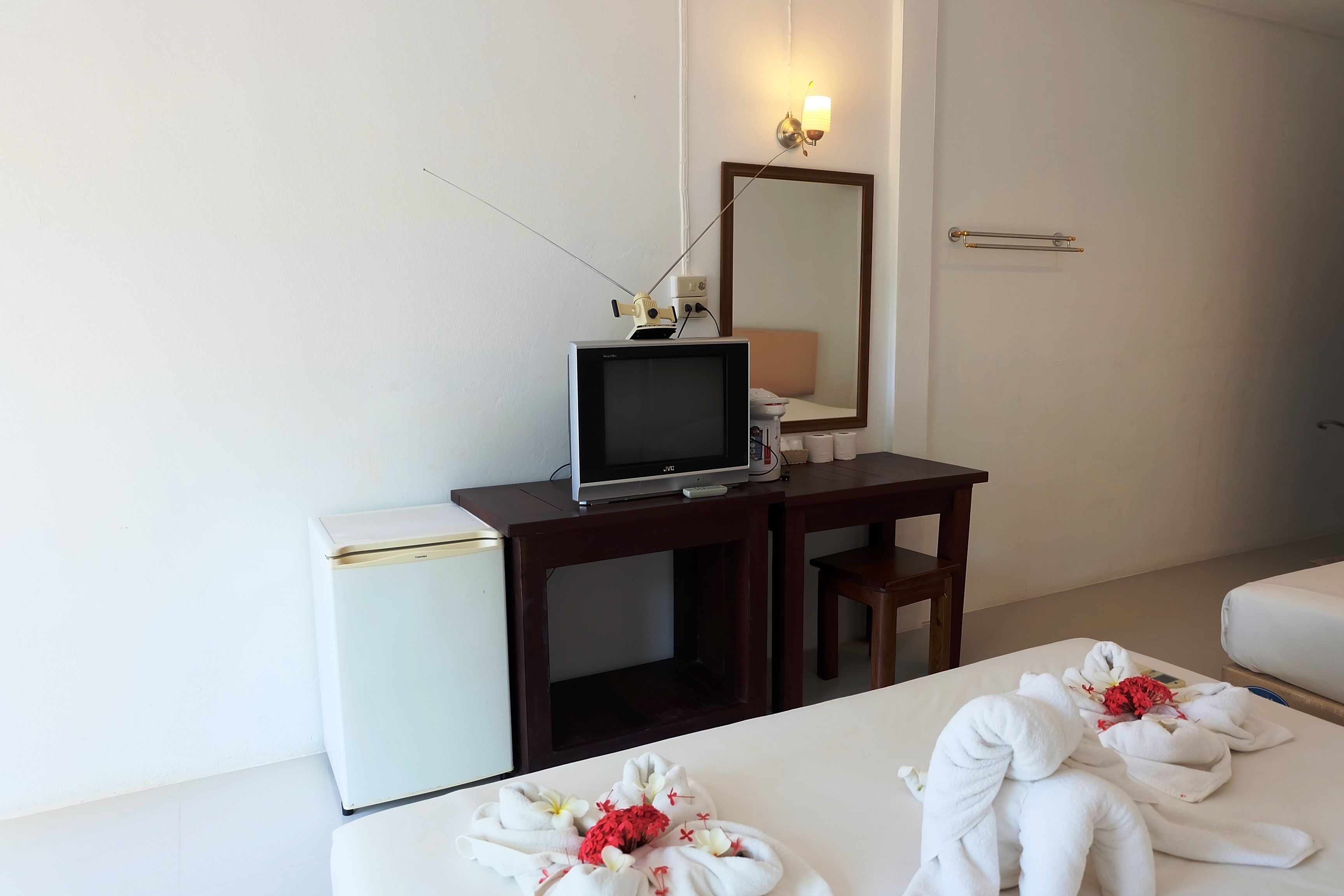 A hotel room with a desk, a television, a small refrigerator, a bed with towels, a mirror, and a bedside table.