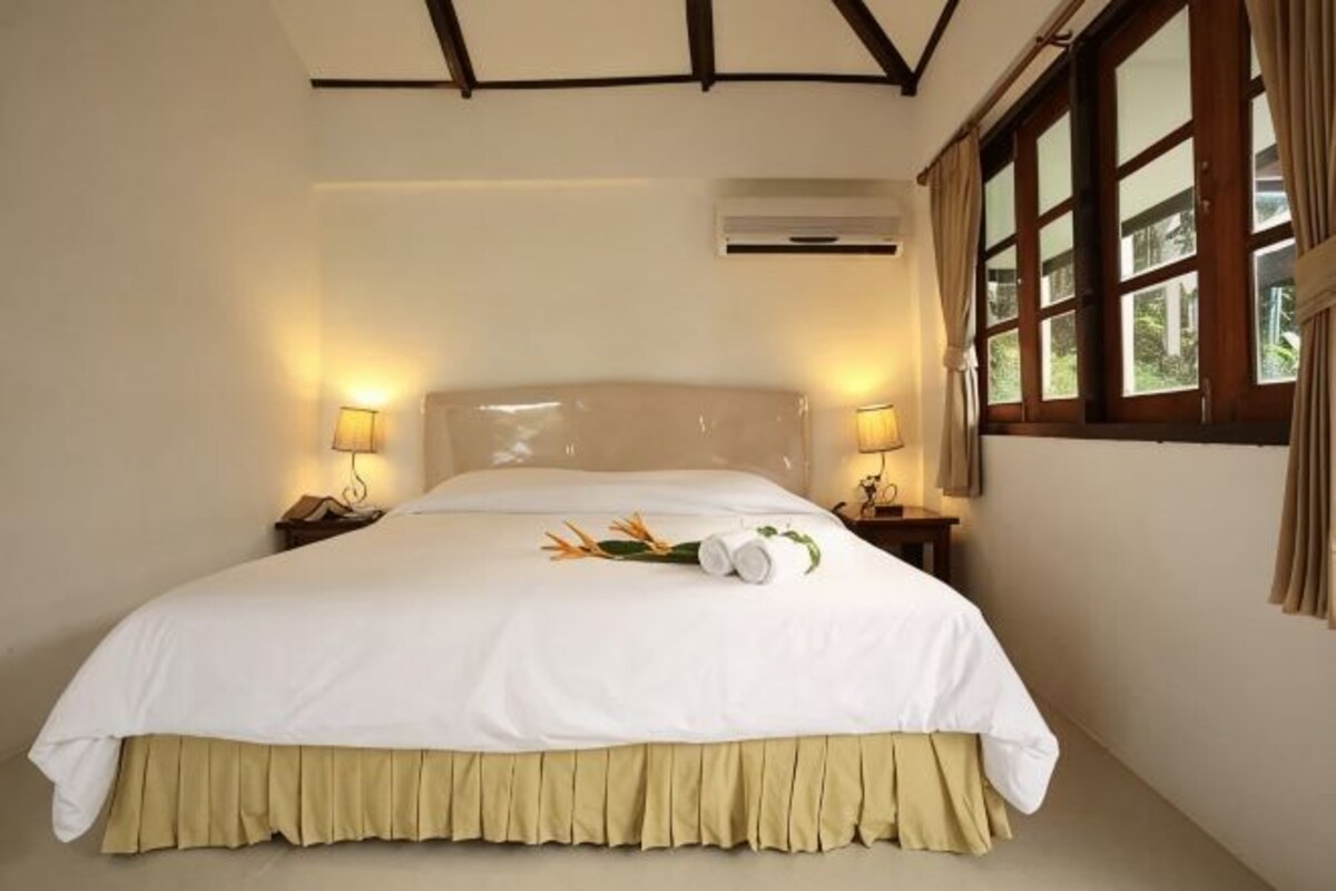 Una cama bien hecha con una colcha blanca y falda color beige, dos lámparas de noche, un equipo de aire acondicionado y una ventana con cortinas.