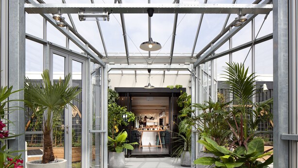 Un invernadero con techo de vidrio, plantas en macetas y vista a una habitación con mesa y sillas.