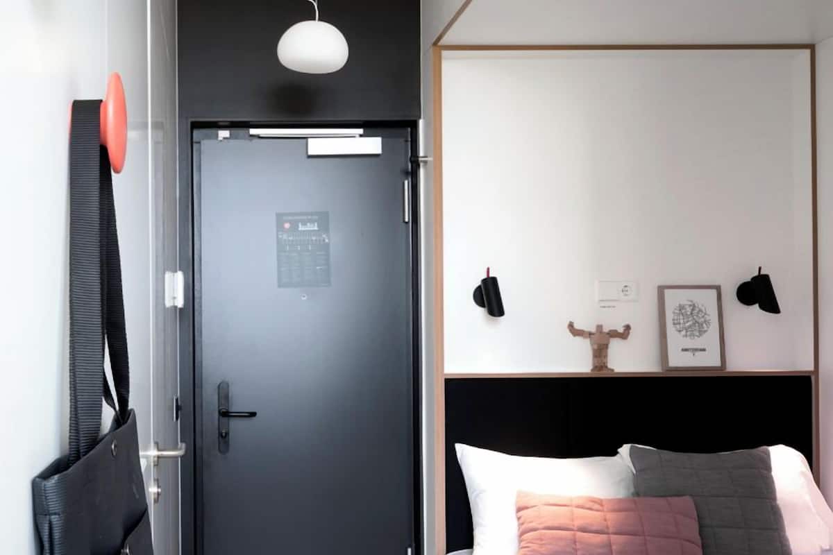 Un dormitorio moderno con una puerta oscura, una cama blanca con muchos almohadones, suelo de madera y un escritorio pequeño con cajones.