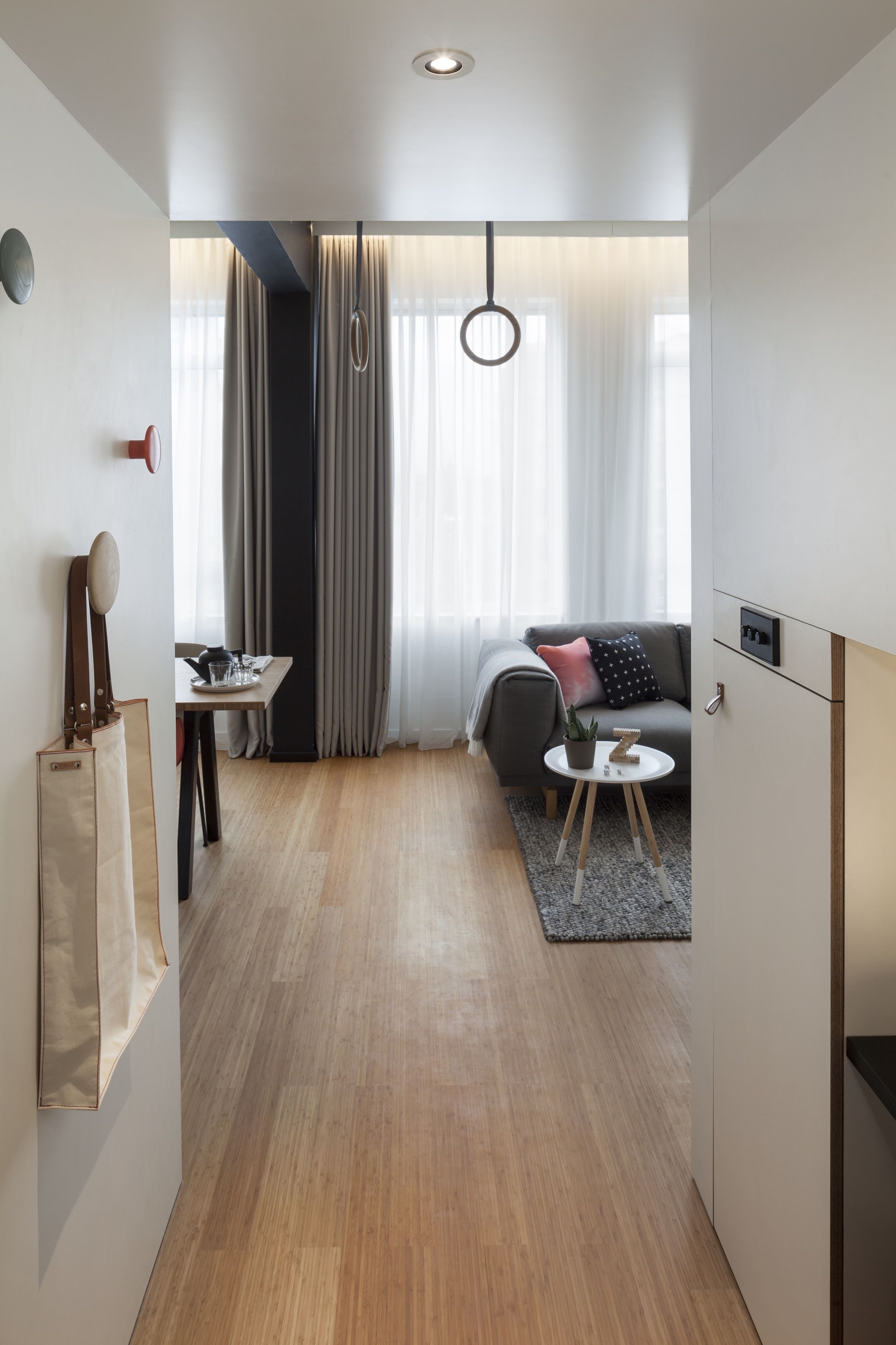 Un espacio habitable moderno y compacto con suelo de madera, un sofá con cojín negro y rosa, una pequeña mesa redonda con una planta y una zona de comedor con mesa y sillas.