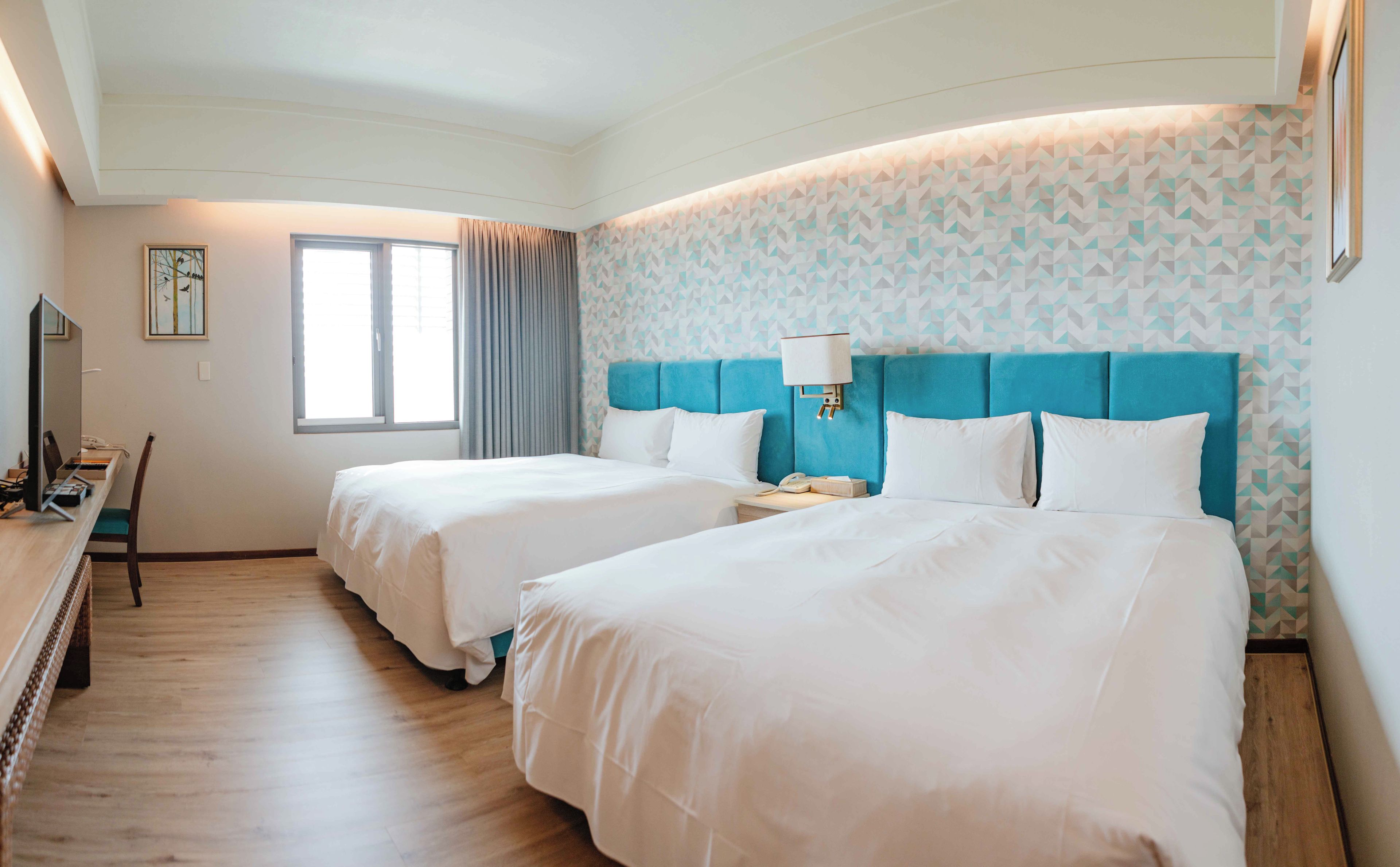 A hotel room with two beds, a desk with a chair, a window with curtains, and a patterned wall.