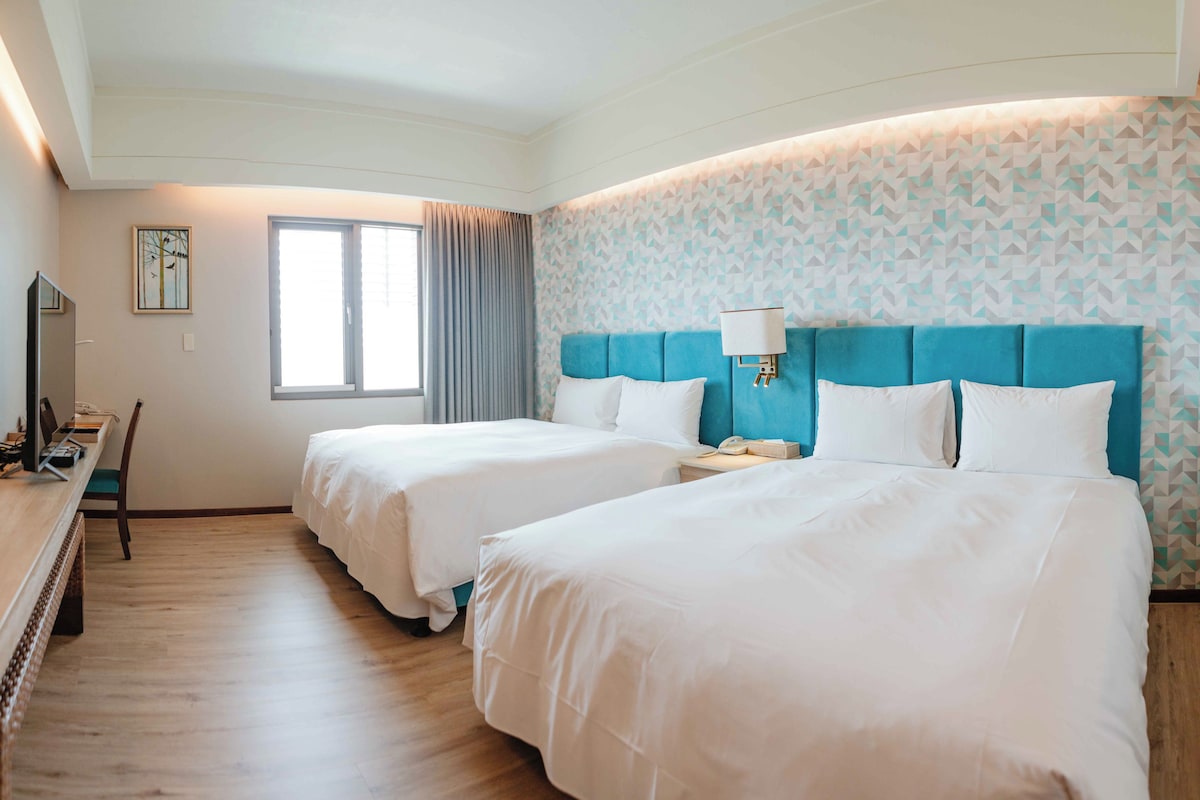 A hotel room with two beds, a desk with a chair, a window with curtains, and a patterned wall.