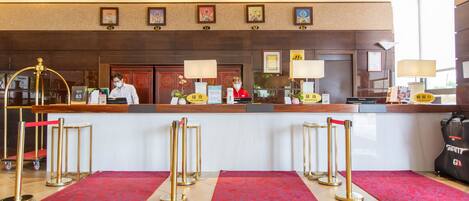 Un'area di reception di un hotel con una scrivania, due receptionist, una cassa registratore e un pavimento ricoperto di tappeto rosso.