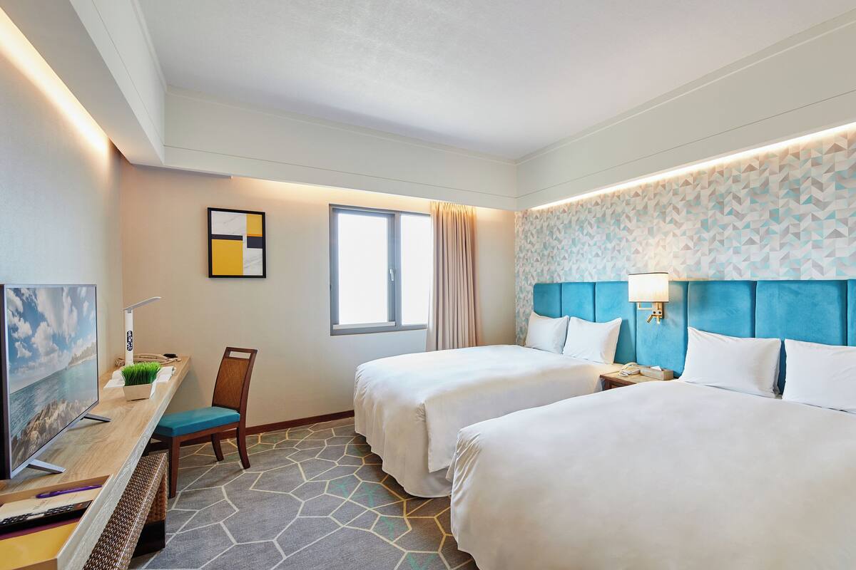 Habitación de hotel con dos camas, un escritorio con computadora, un televisor y una ventana con cortinas.