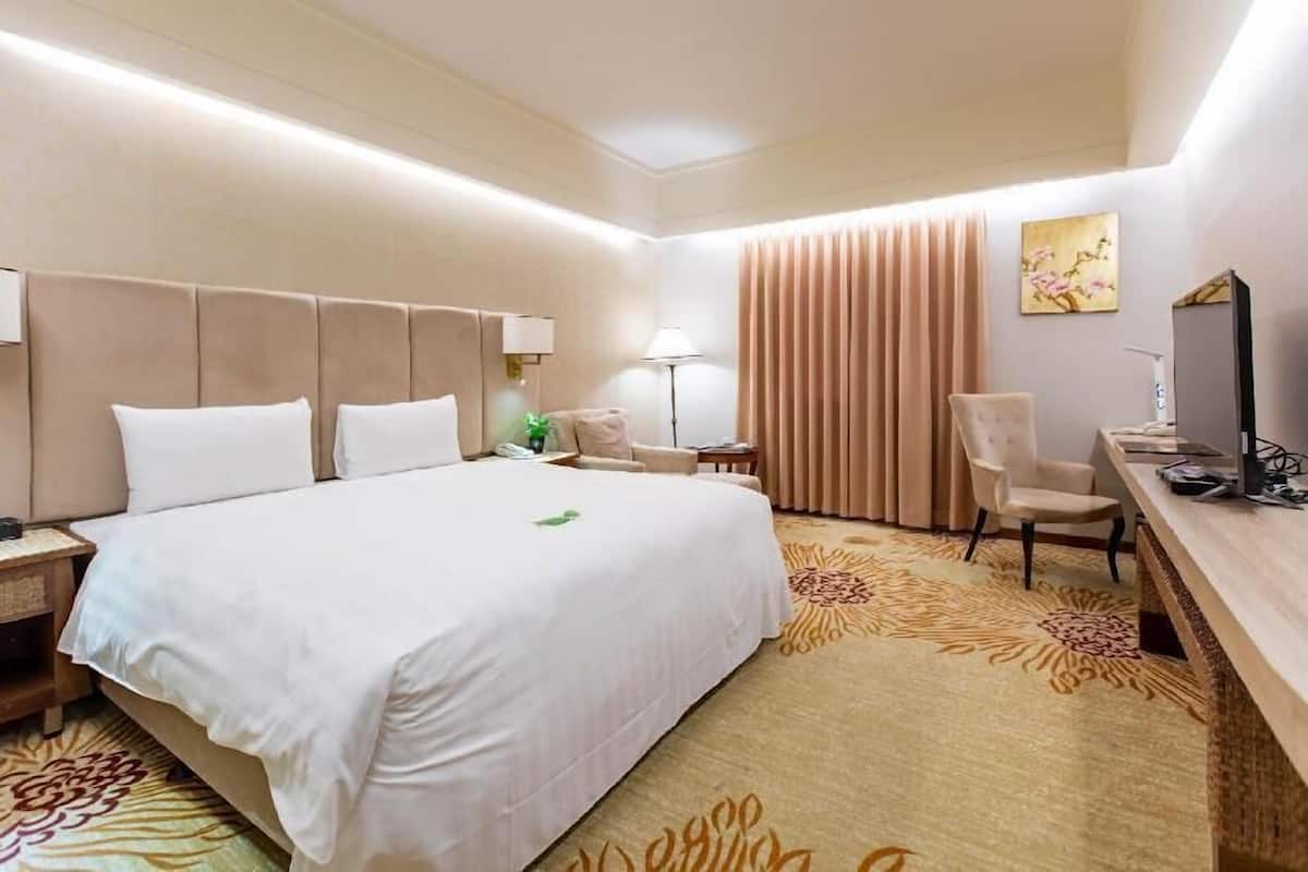 Habitación de hotel con una cama grande, un escritorio, una silla, una lámpara, un cuadro y una alfombra.