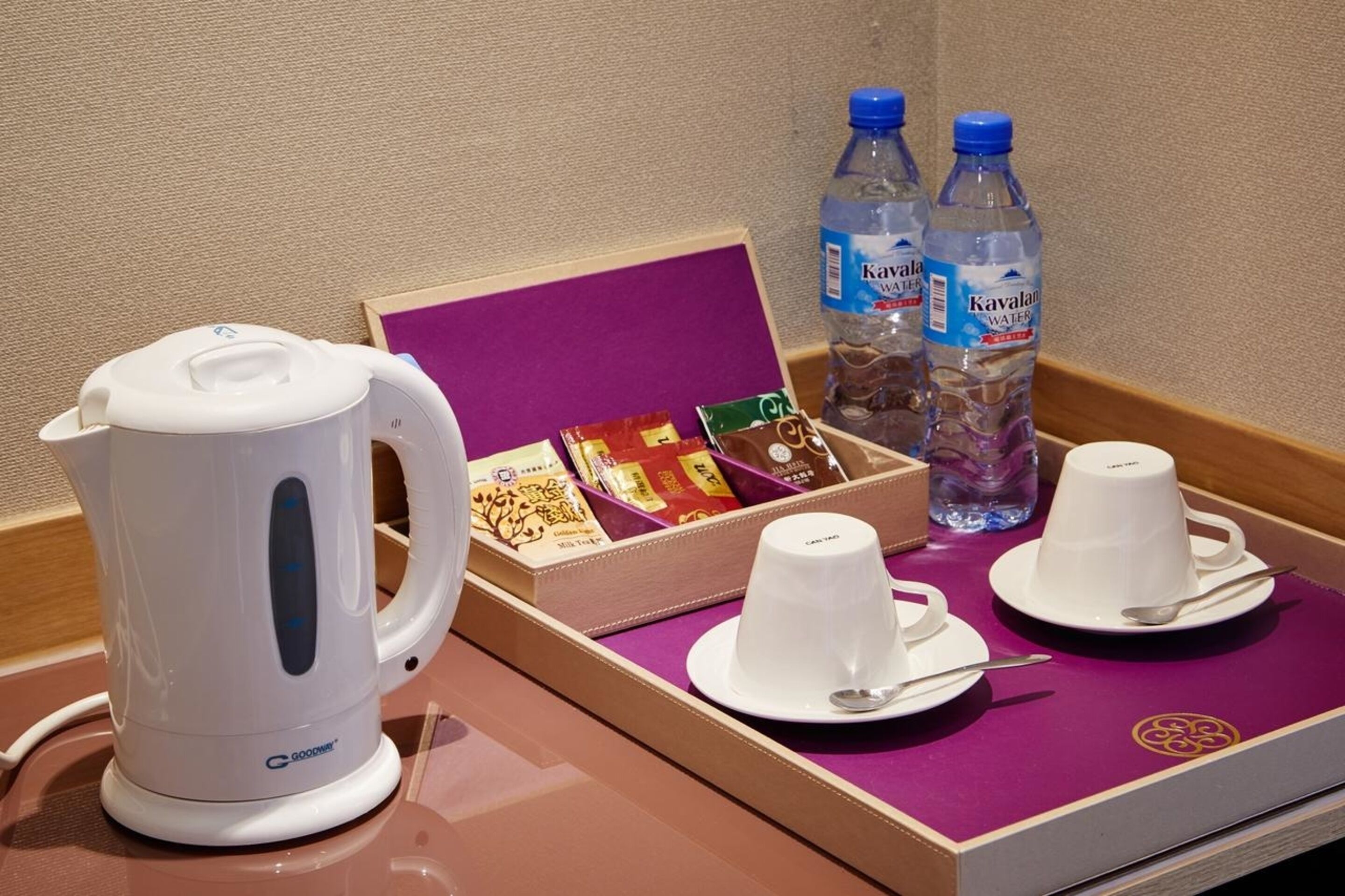 A hotel room with a white electric kettle, a tray with two cups and spoons, a box of tea bags, and two bottles of water.