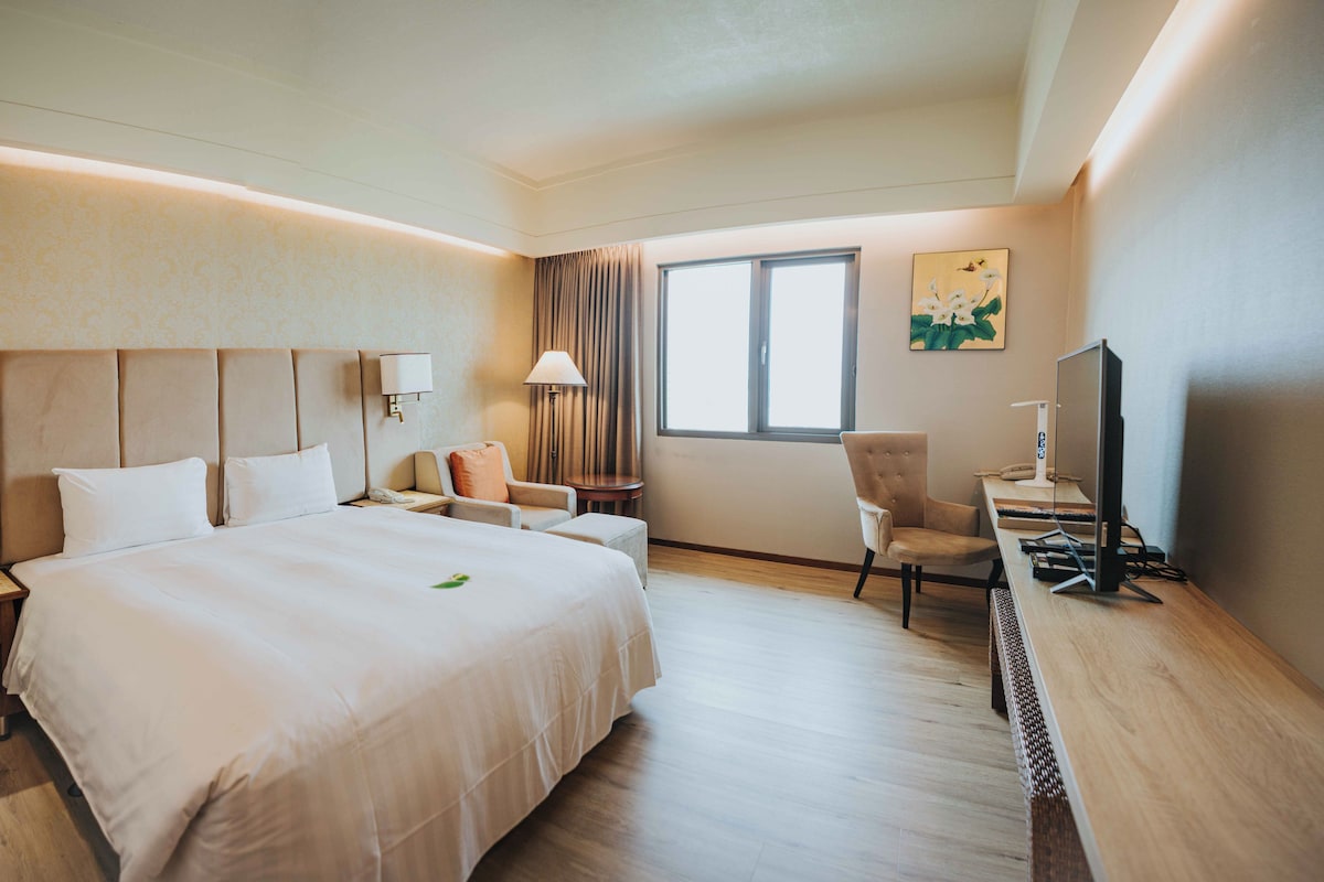 A hotel room with a large bed, a desk with a computer, a chair, and a window with curtains.