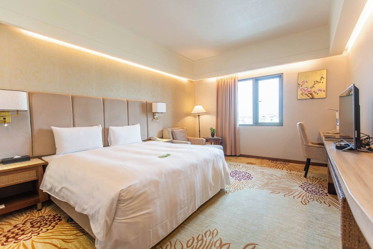 Una habitación de hotel con una cama grande, lámparas de noche, un escritorio con una computadora, una silla y una ventana con cortinas.