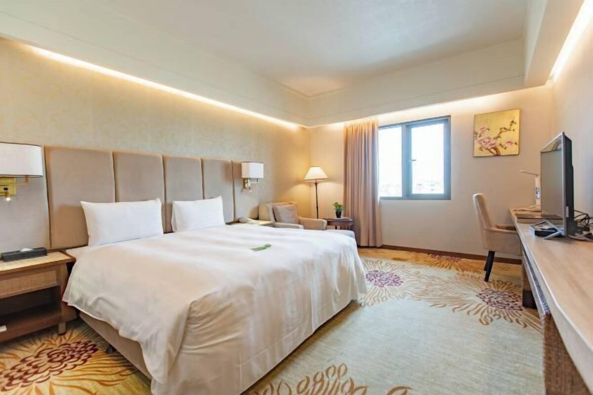 Habitación de hotel con una cama grande, mesitas de noche, un escritorio, una silla, un televisor y una ventana con cortinas.