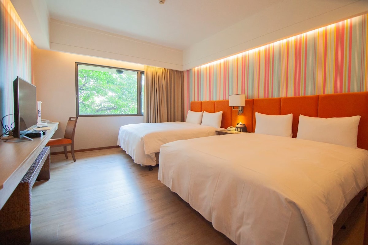 A hotel room with two beds, a desk with a computer, a window with curtains, and a striped wall.