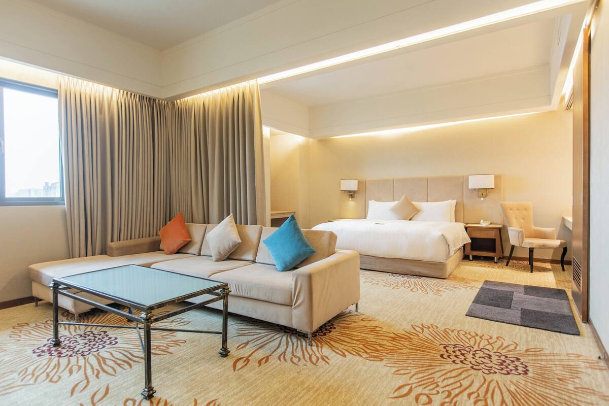 Habitación de hotel con sofá, cama, mesita de noche, lámpara y ventana con cortinas.