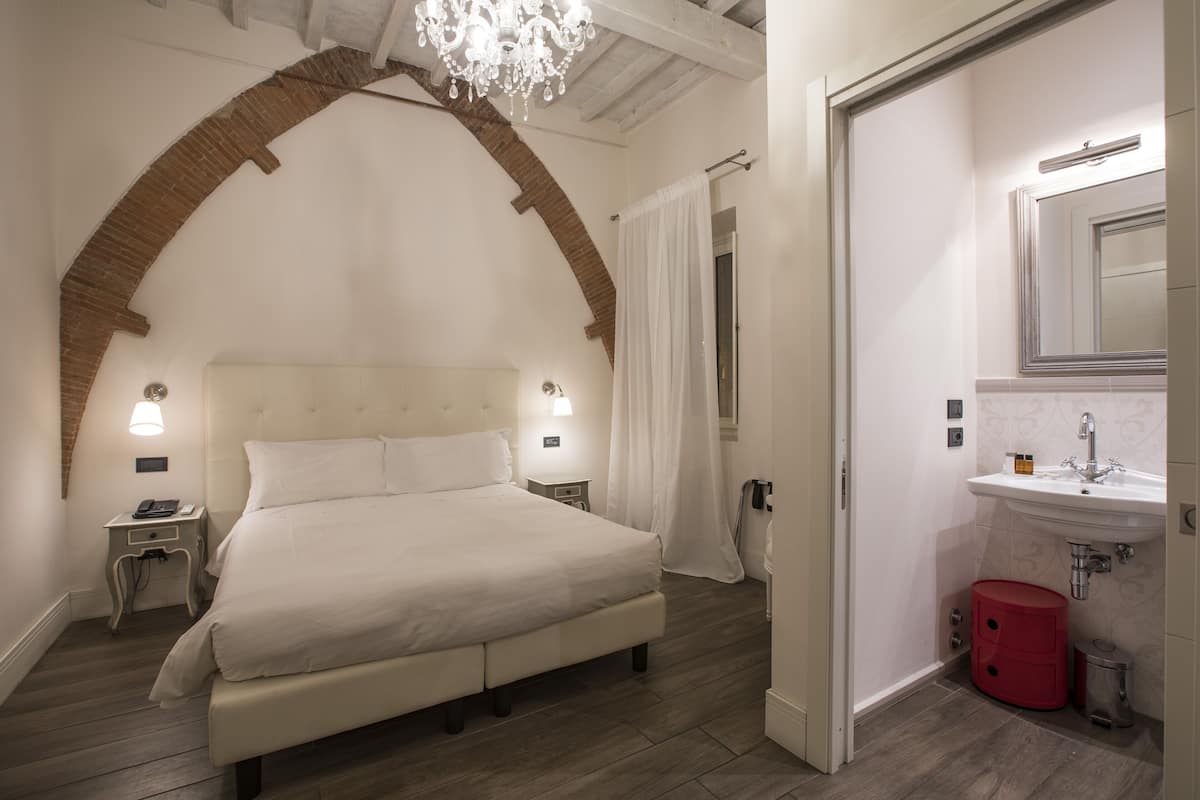A bedroom with a large bed, a nightstand, a lamp, a chandelier, and a bathroom with a sink and mirror.