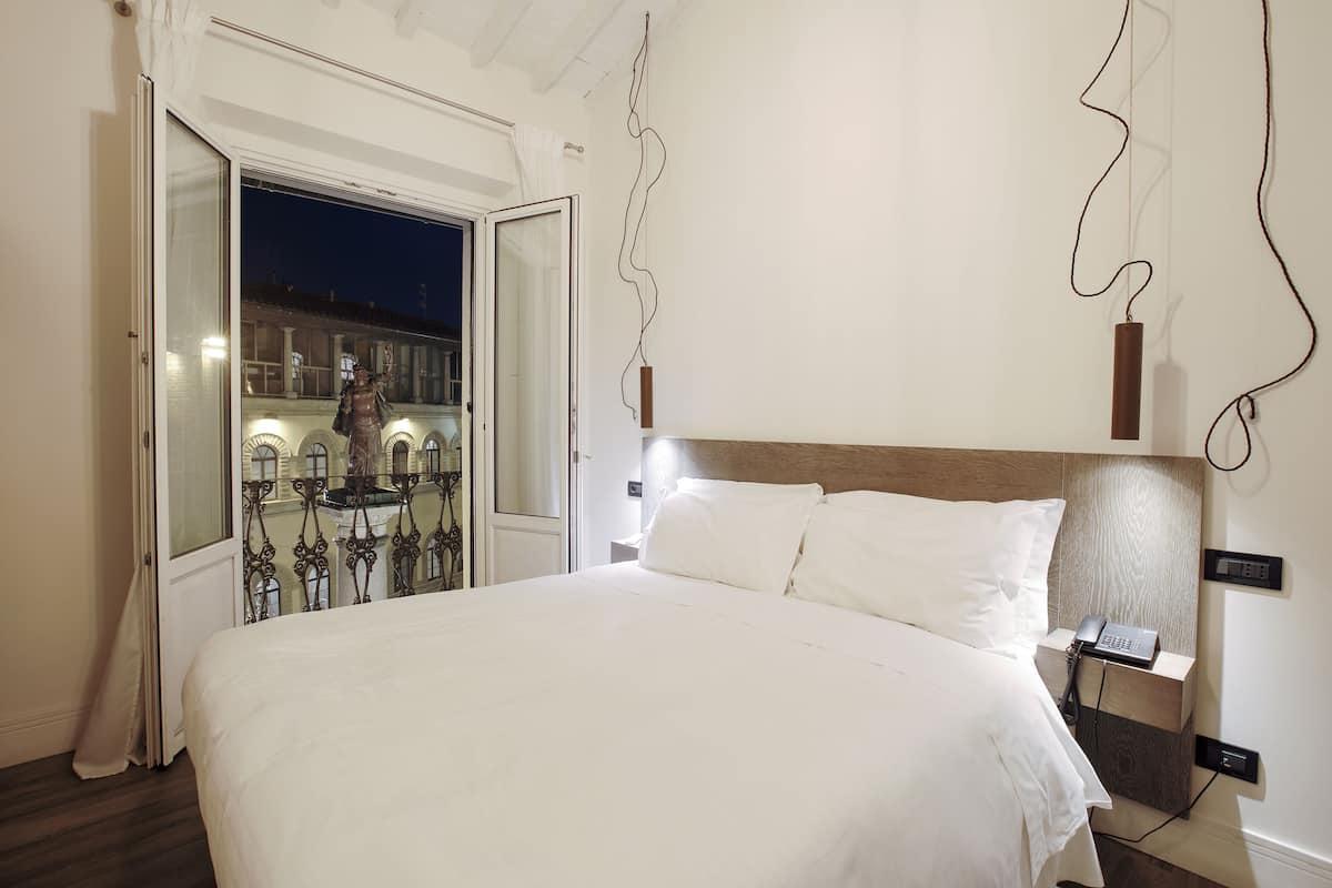 A hotel room with a large bed, a bedside table with a phone, a wall-mounted light, and a window with a view of buildings outside.