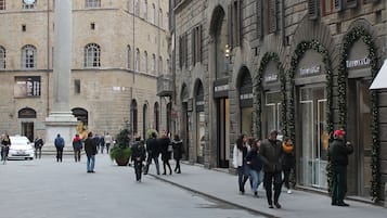 Una plaza histórica con un monumento central, edificios de piedra con ventanales arqueados y algunas personas caminando.