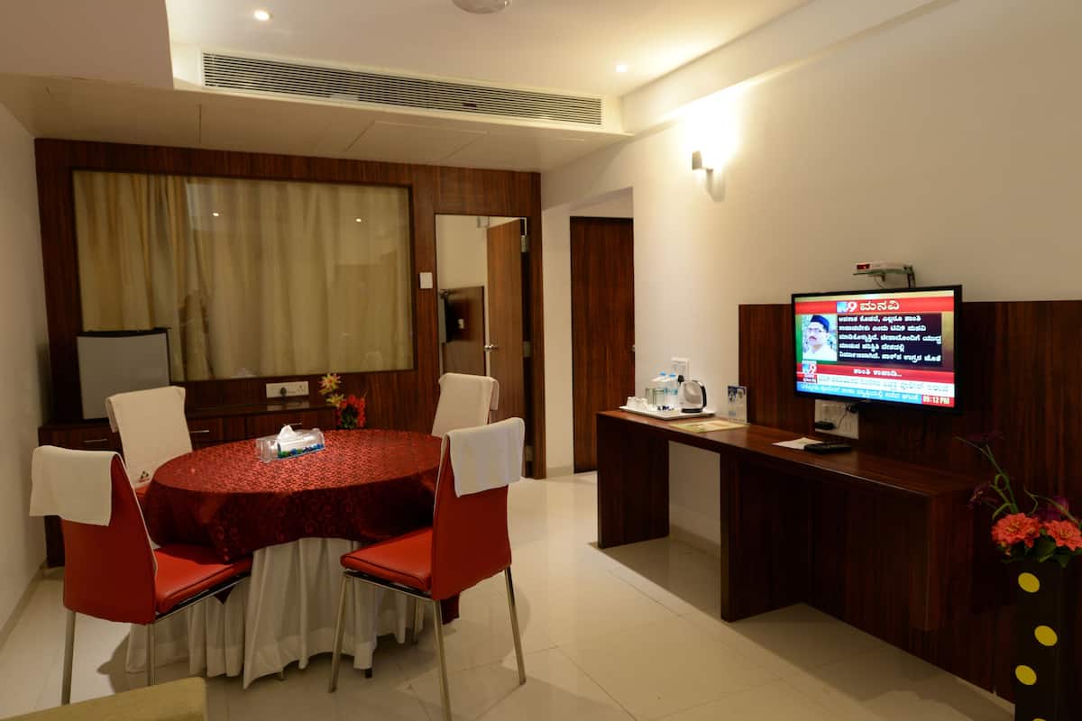 A hotel room with a round dining table, red chairs, a wooden reception desk, and a television displaying a news channel.