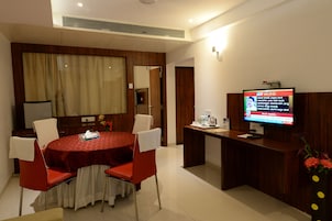 A hotel room with a round dining table, red chairs, a wooden reception desk, and a television displaying a news channel.