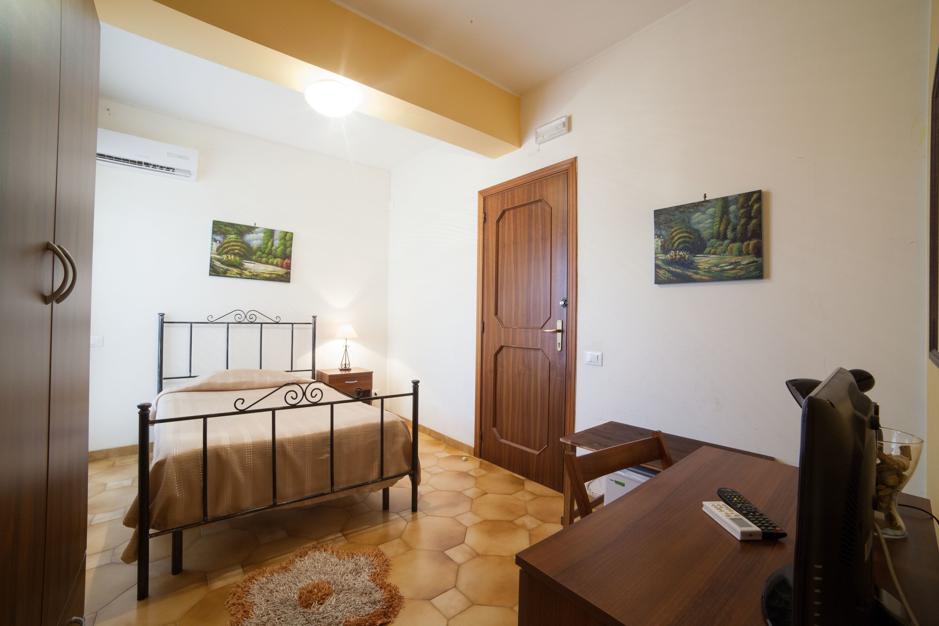 Un dormitorio con cama, mesita de noche, lámpara y un escritorio con monitor de computadora.