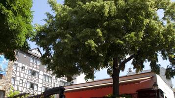 A cobblestone plaza with a large tree, a building with a red awning, and a stone wall.
