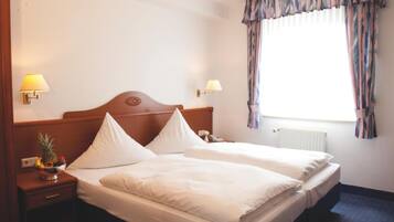 A double bed with two pillows, a wooden headboard, a nightstand with a lamp, a fruit bowl, and a window with curtains.