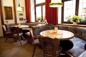A restaurant interior with round tables and booths, wooden chairs, and potted plants by the windows.