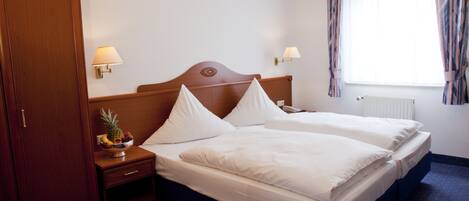 A hotel room with two single beds, a wooden wardrobe, bedside lamps, a fruit bowl, and a window with curtains.
