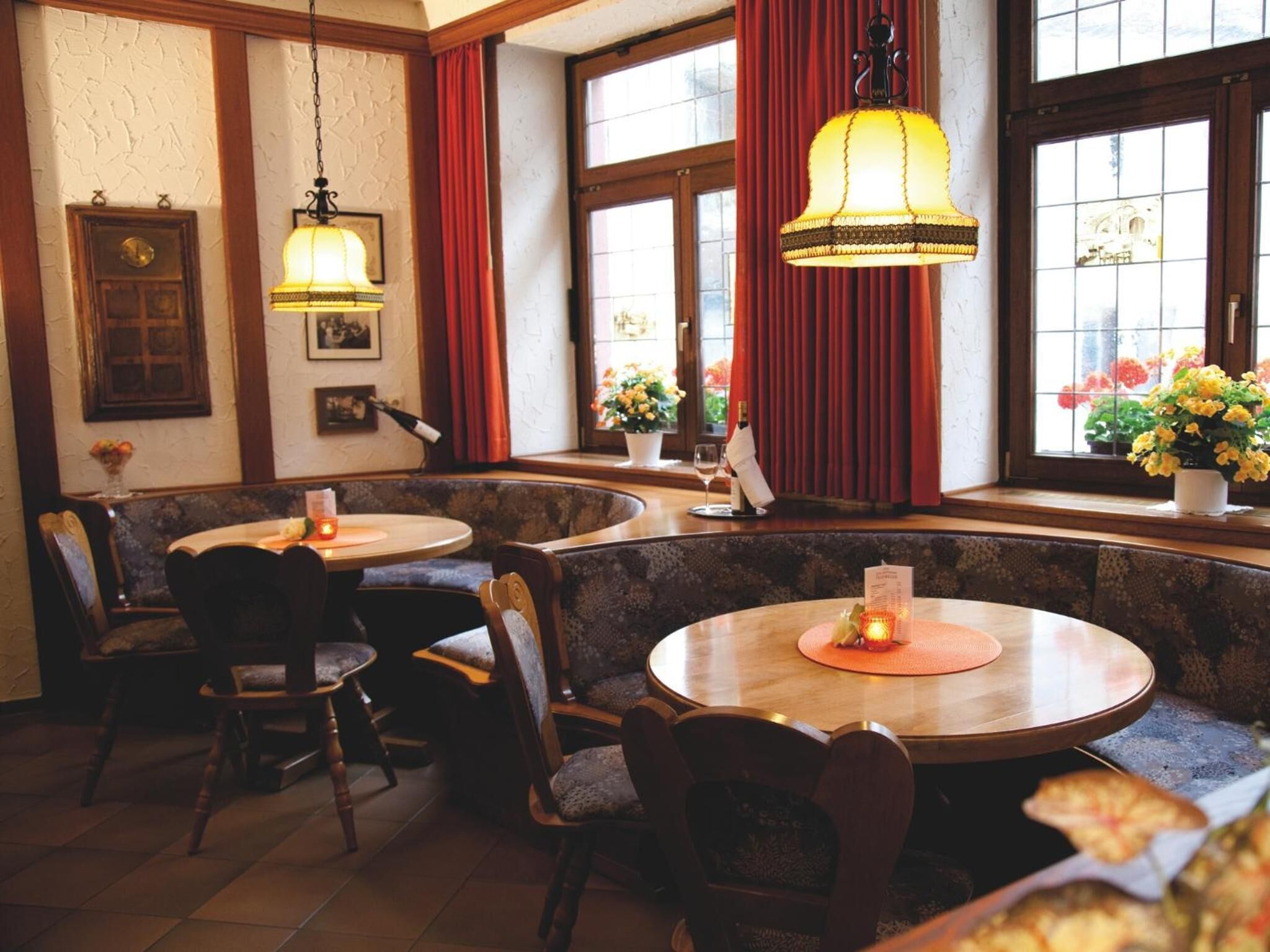 A cozy restaurant interior with round tables and booths, warm lighting, and large windows with curtains.