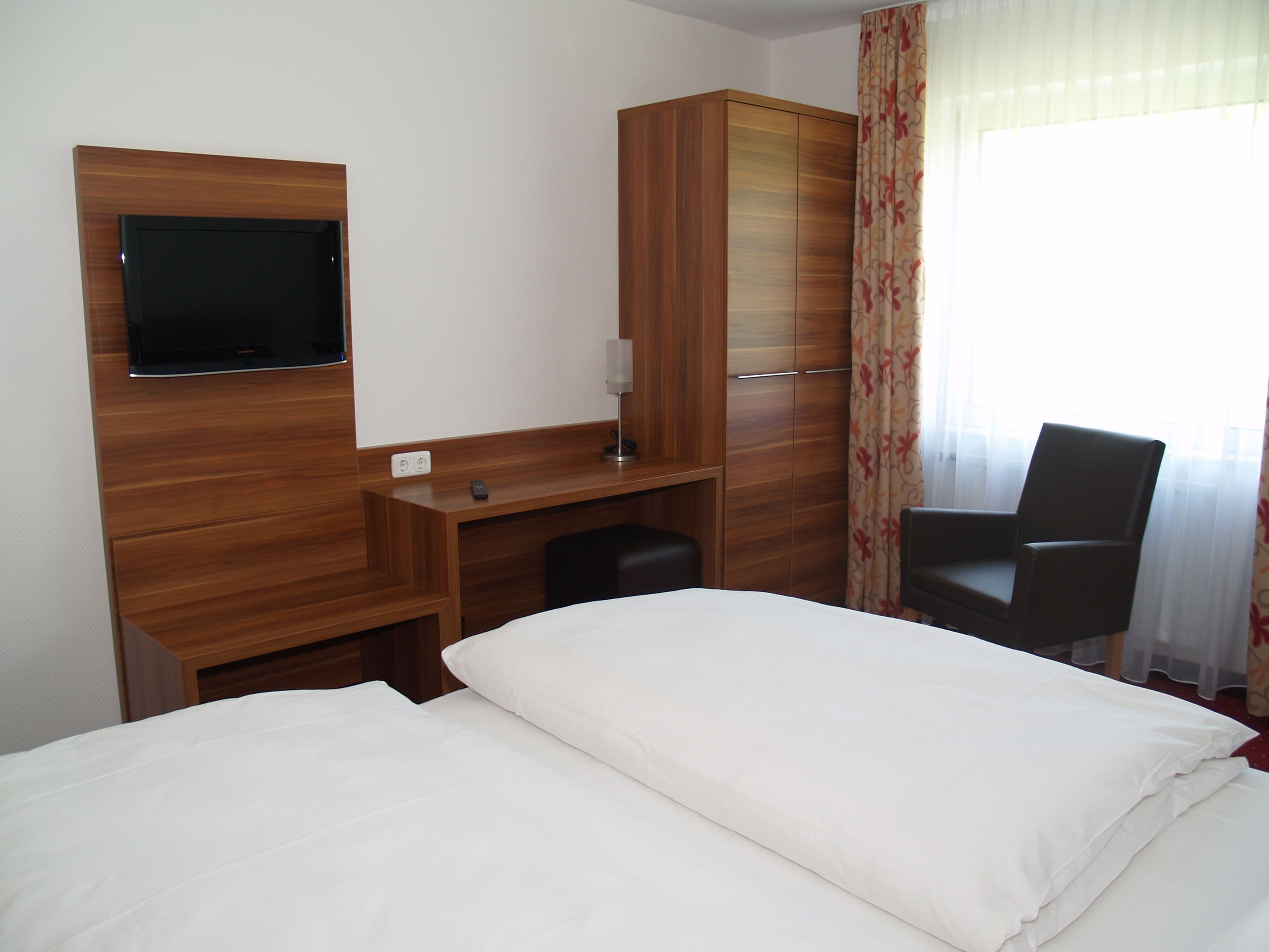 A hotel room with a bed, a desk, a chair, a TV mounted on the wall, and a wardrobe.