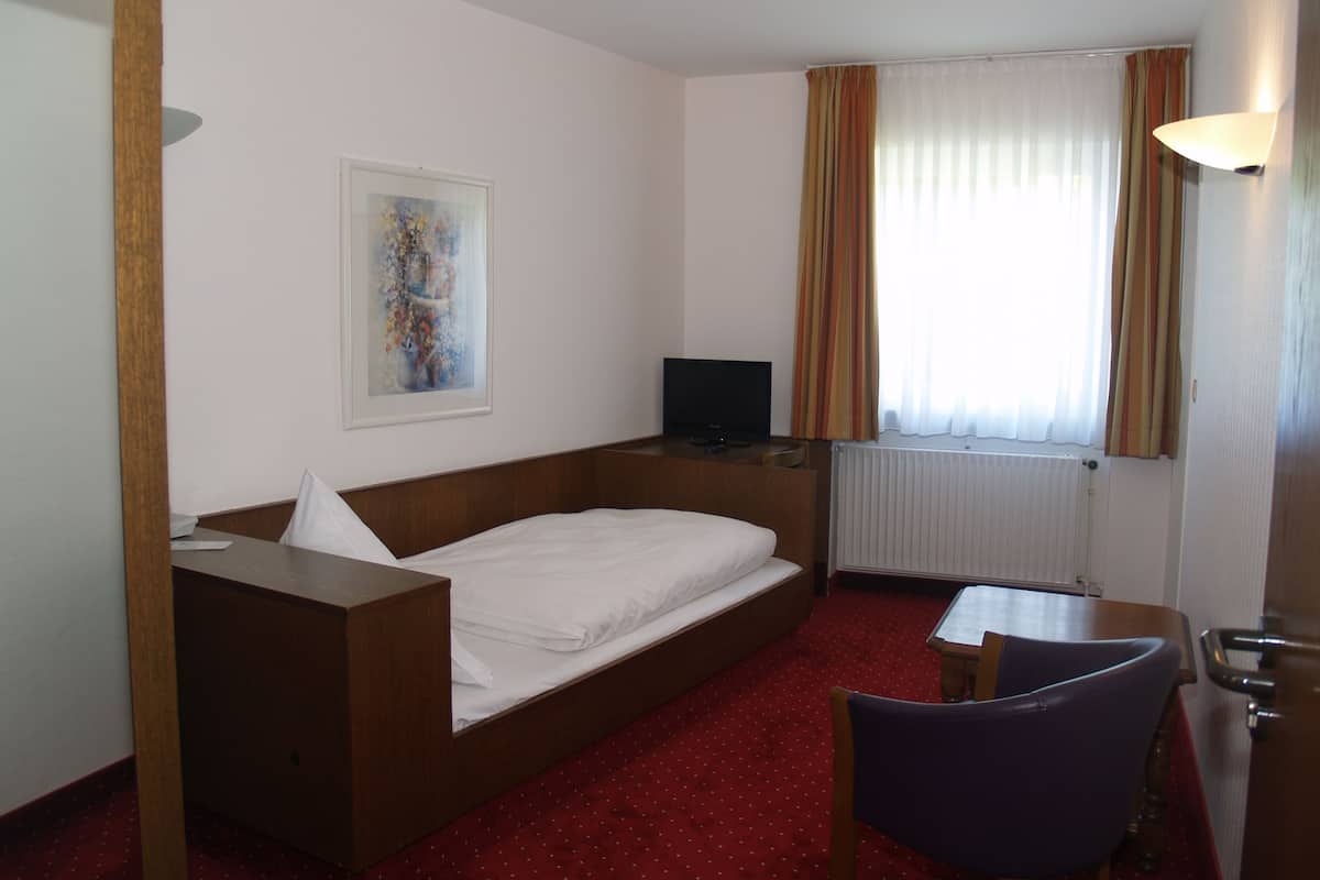 Une chambre d’hôtel avec un lit simple, un bureau avec une chaise, une télévision et une fenêtre avec des rideaux.