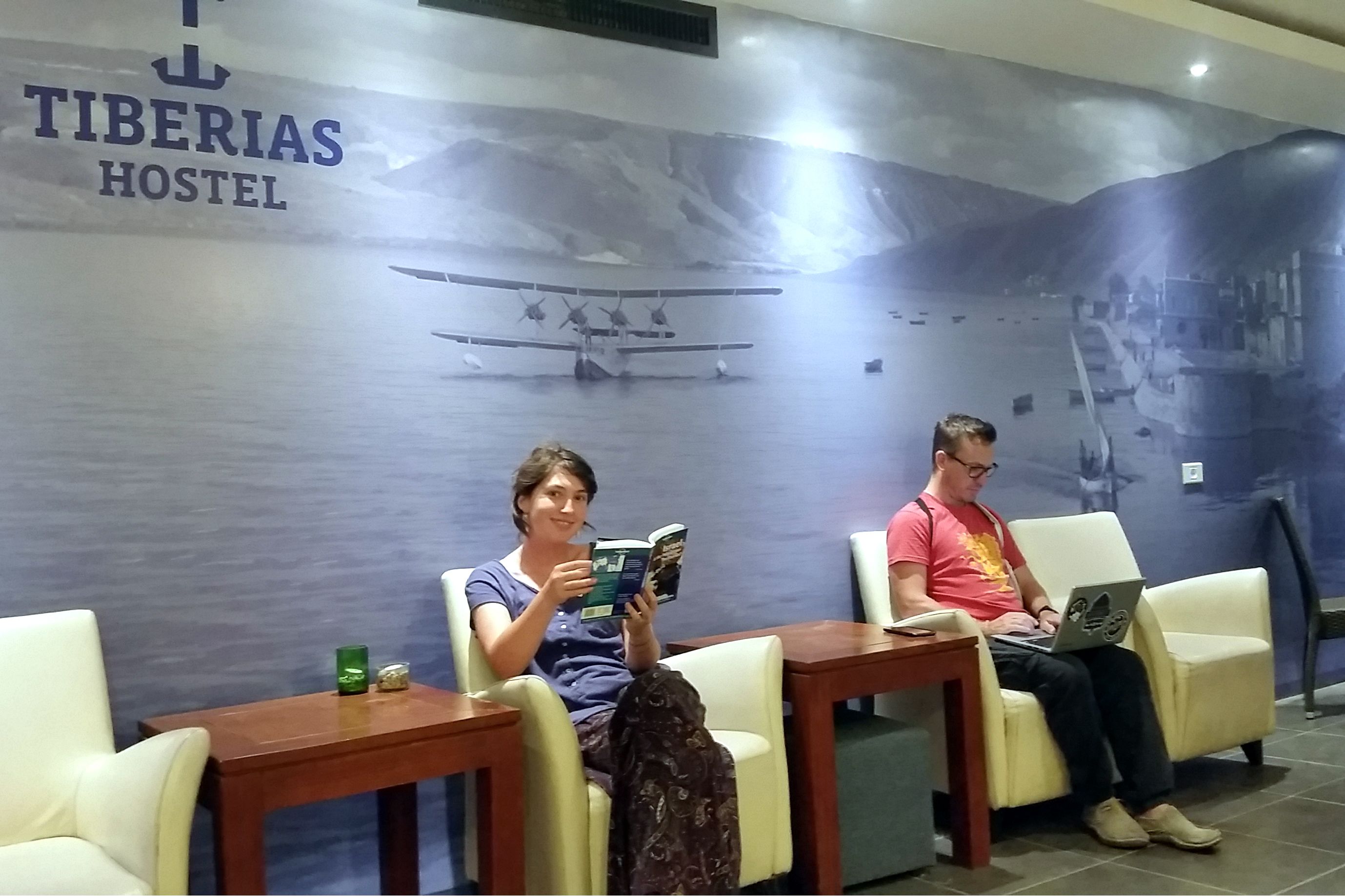 A hostel interior with seating areas, a mural of an airplane, and a sign reading "TIBERIAS HOSTEL".