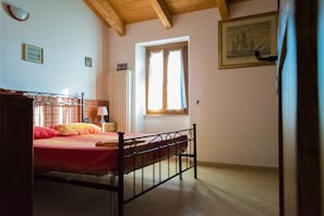 A bedroom with a metal bed frame, a wooden headboard, a window with curtains, a radiator, and a framed picture on the wall. - Luce Stellata (Podenzana)