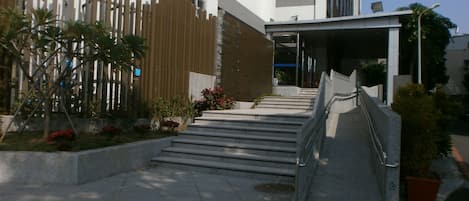 Entrada de un edificio con una escalera que lleva a un área cubierta, rodeada de plantas en macetas y una valla de madera.