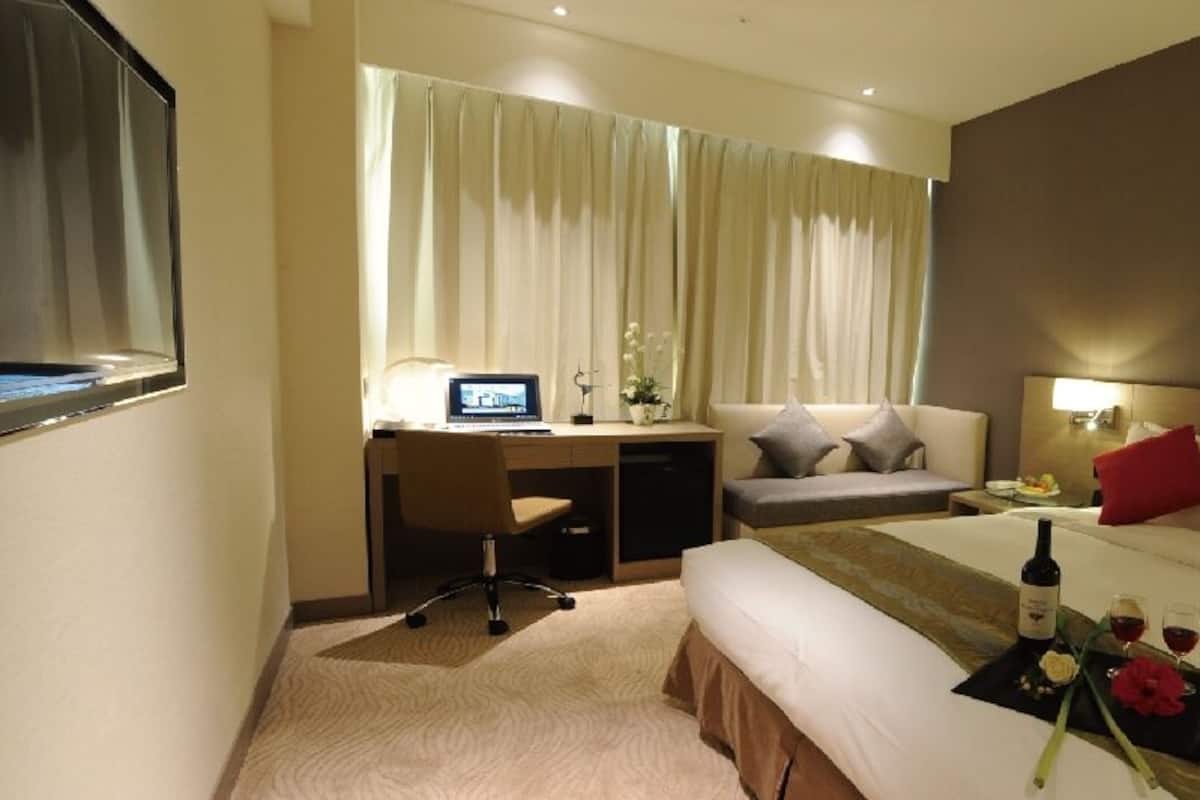 A hotel room with a bed, a desk, a chair, a small table with a bottle, and a telephone.