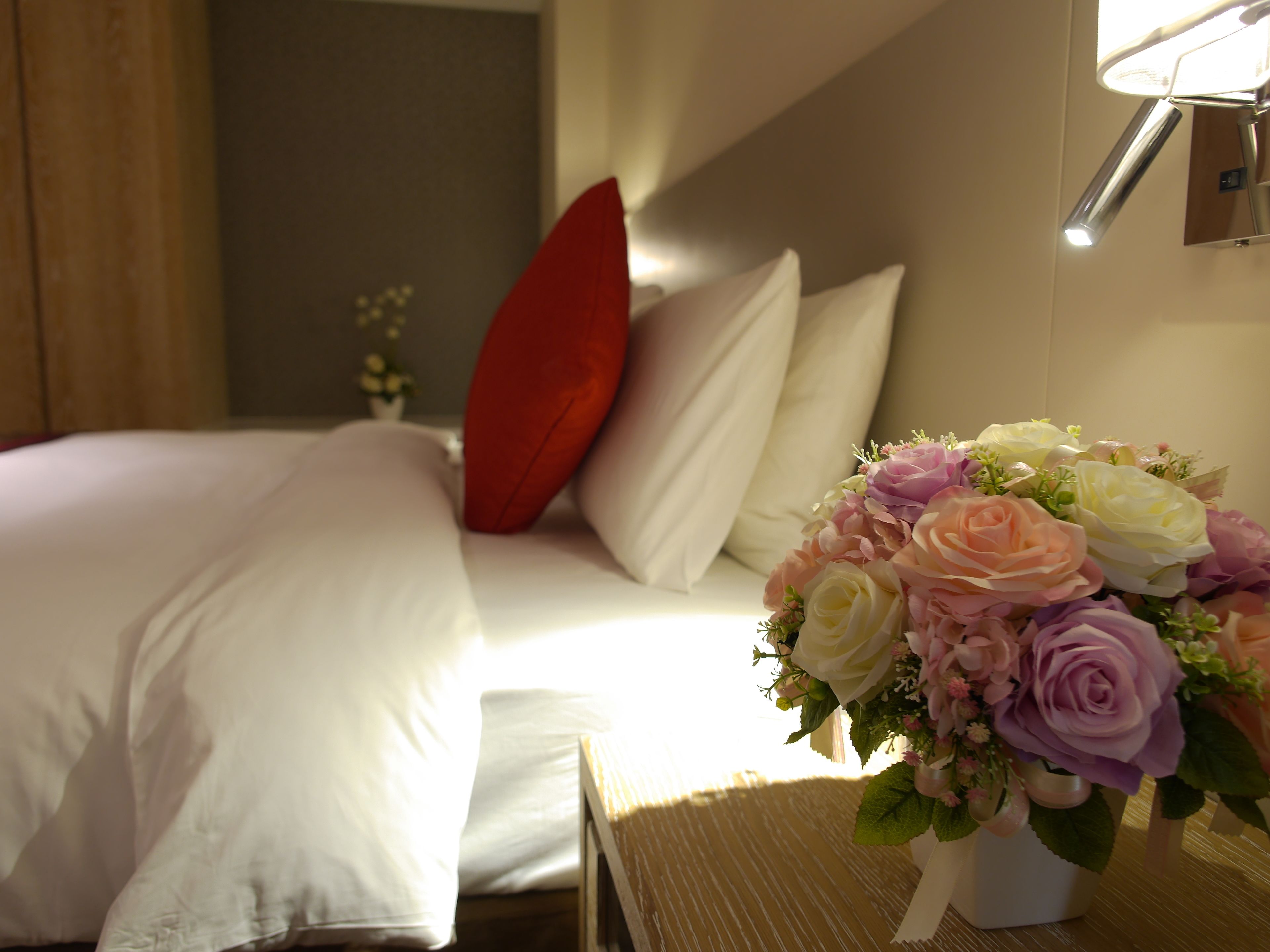 Et hotelværelse med en pænt redt seng, en rød pude og en blomstrende buket på sengebordet.