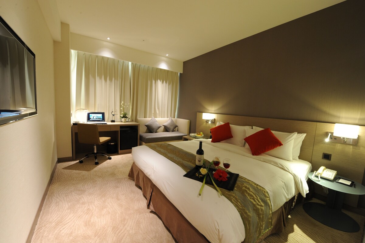 A hotel room with a bed, desk, and chair. There is a television on the wall, a window with curtains, and a bedside table with a phone and a lamp.
