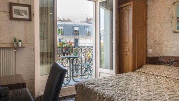 A hotel room with a bed, a desk with a chair, a radiator, and a balcony with a view of buildings. - Familia Hotel (Paris)