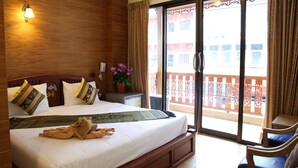 Habitación de hotel con una cama grande, cabecera de madera, mesita de noche con lámpara, una pequeña mesa con una planta y un balcón con vistas a edificios.