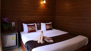Habitación de hotel con pared de madera, cama con ropa blanca, almohadas rojas y doradas, una mesita de noche pequeña con un teléfono y una planta, y un equipo de aire acondicionado sobre la cama.