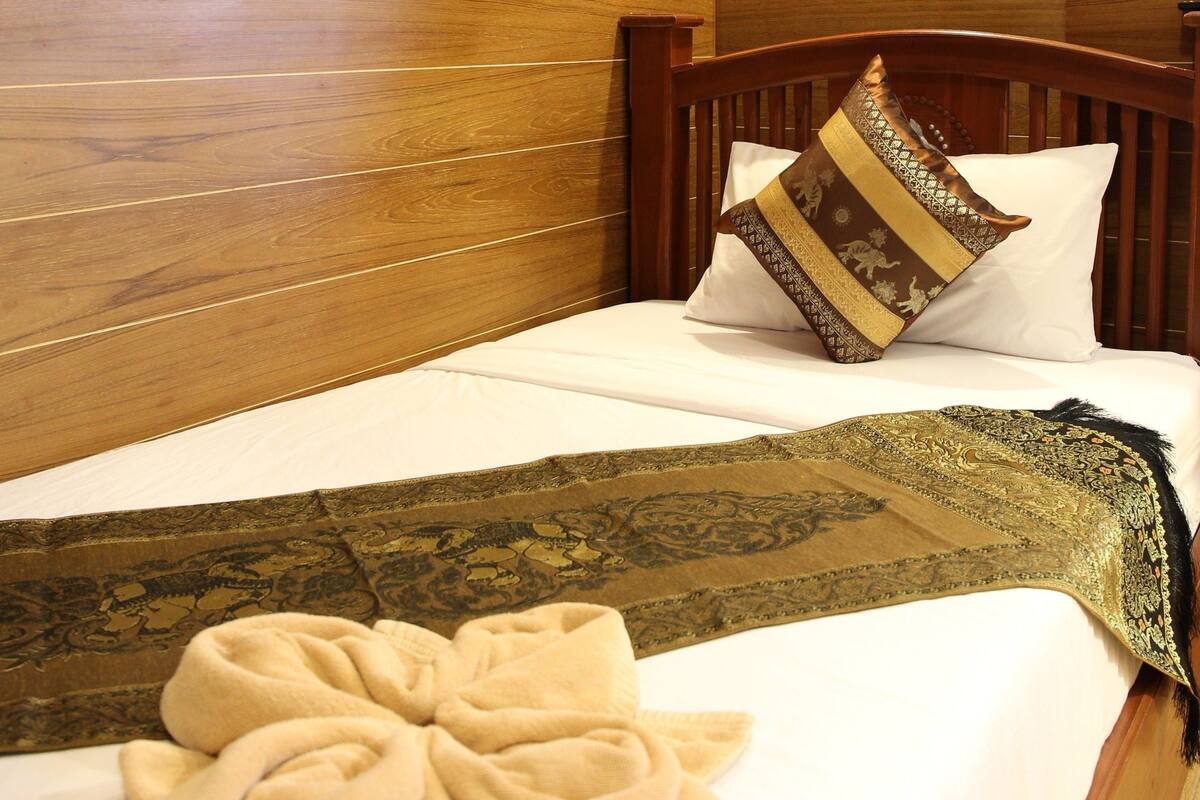 Una cama bien tendida con un plaid decorativo y una almohada, frente a una pared de madera con una lámpara.