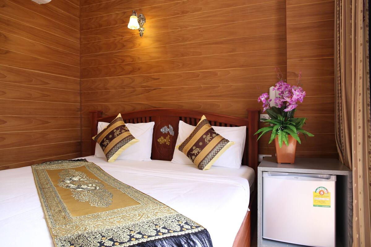 Habitación con paneles de madera, una cama, almohadas, una mesita de noche, una planta en maceta y un refrigerador pequeño.