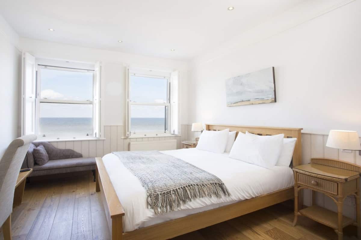 Un dormitorio con una cama grande, dos ventanas con vistas al océano, una mesita de noche con lámpara y una silla.