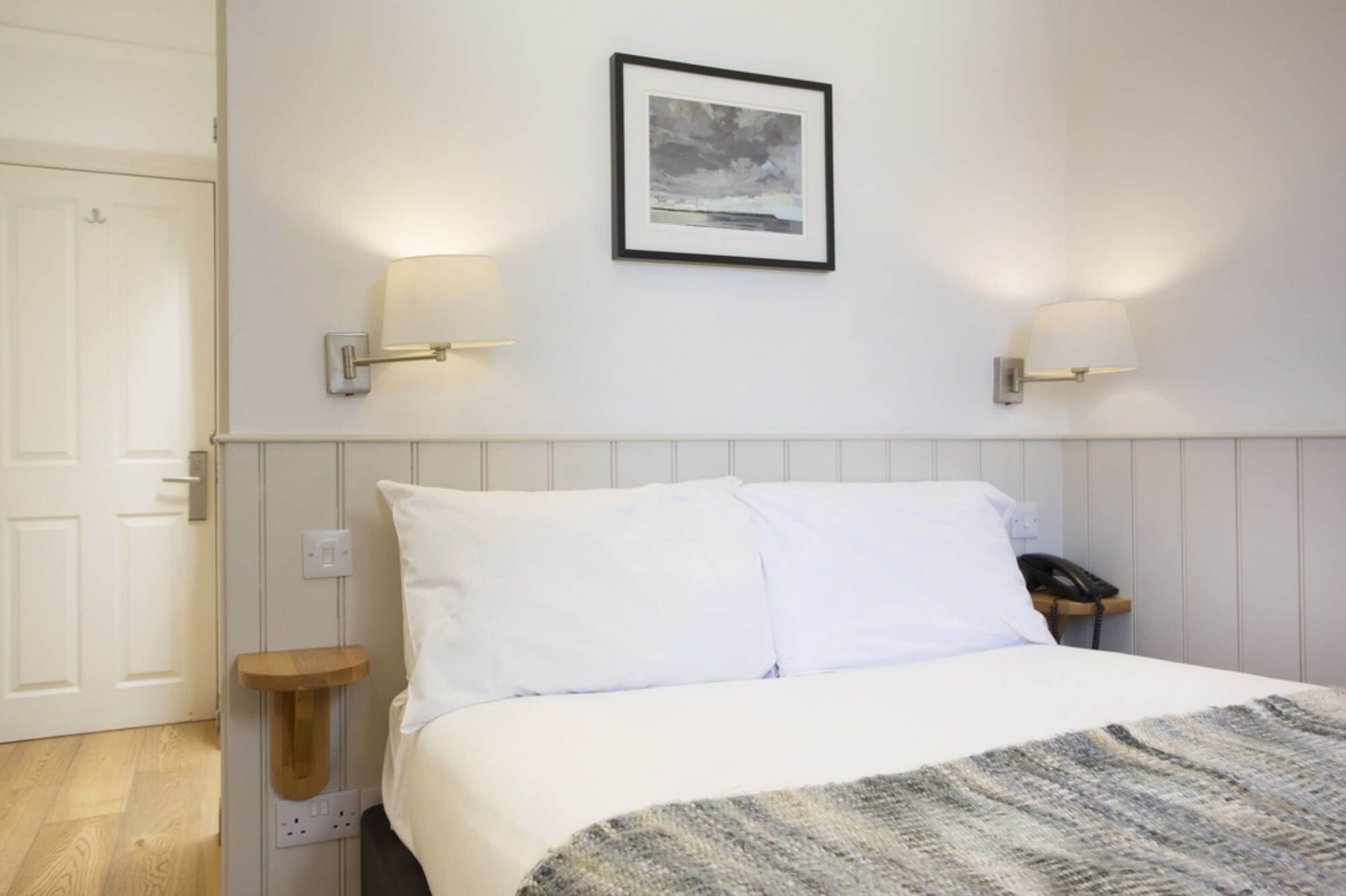 A neatly made bed with white linens, a framed picture on the wall, and two wall-mounted lamps.