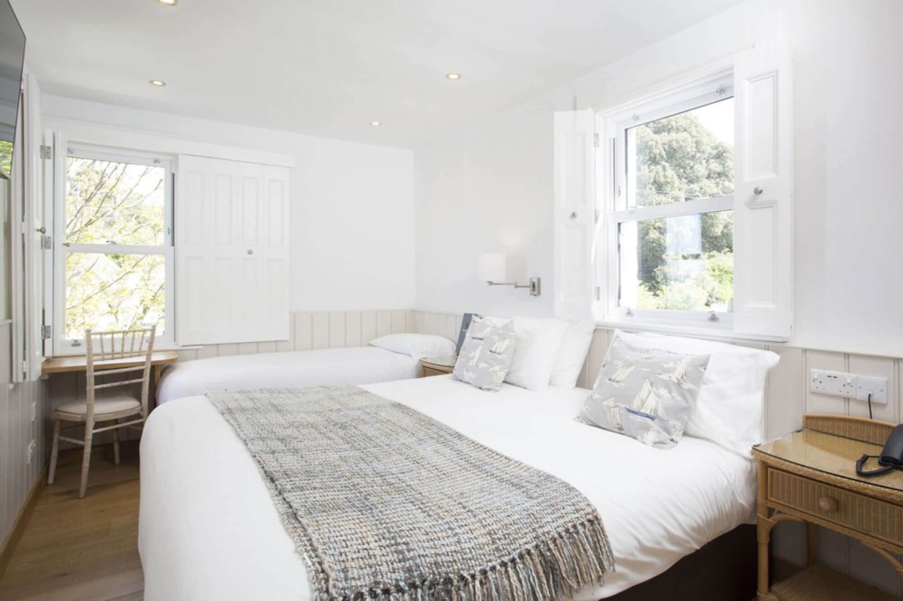 A bedroom with two beds, a desk, a chair, a nightstand, and a window with a view of greenery.